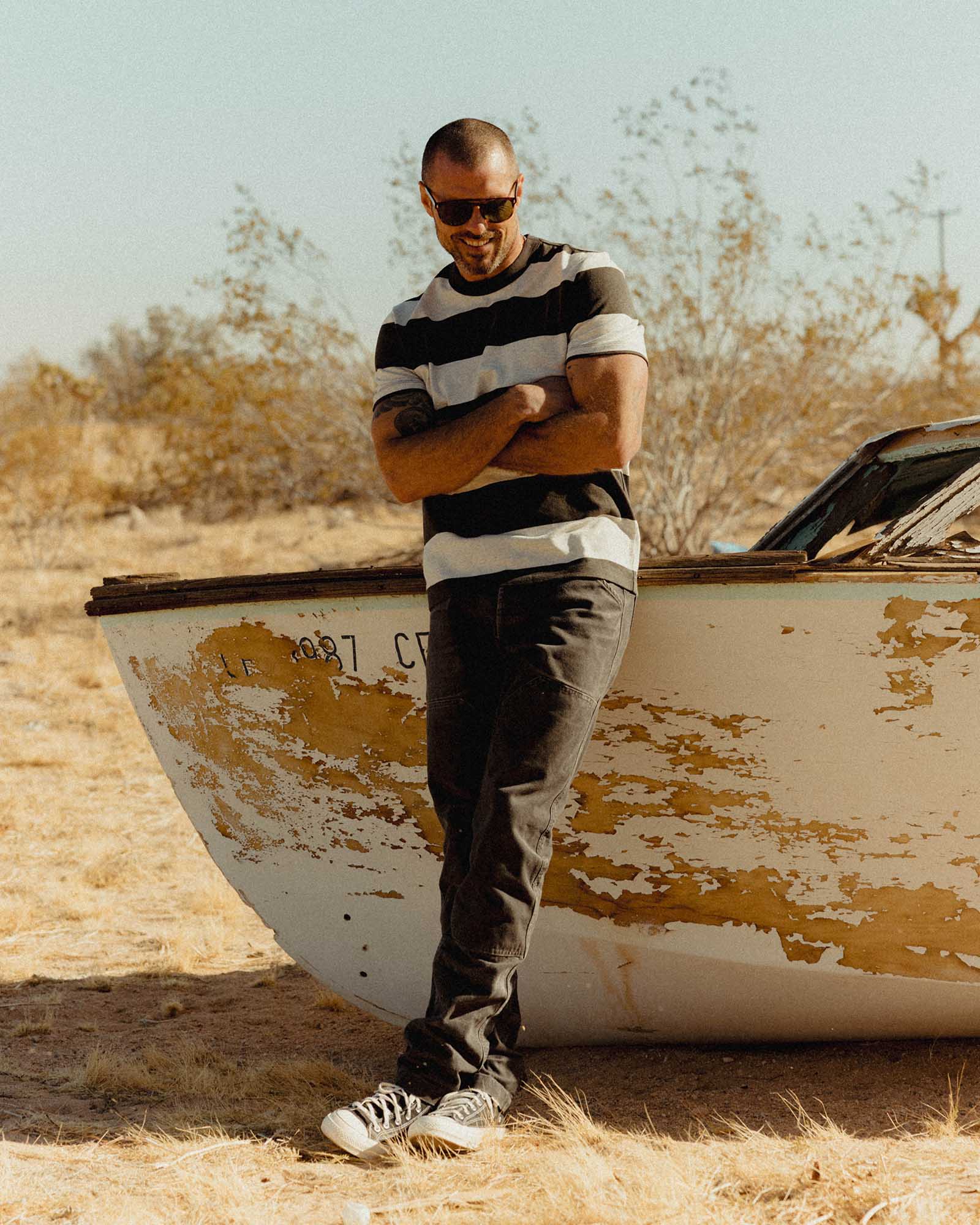 fit model leaning against a boat wearing The Organic Cotton Shop Tee in Faded Black Stripe, Knits by Taylor Stitch